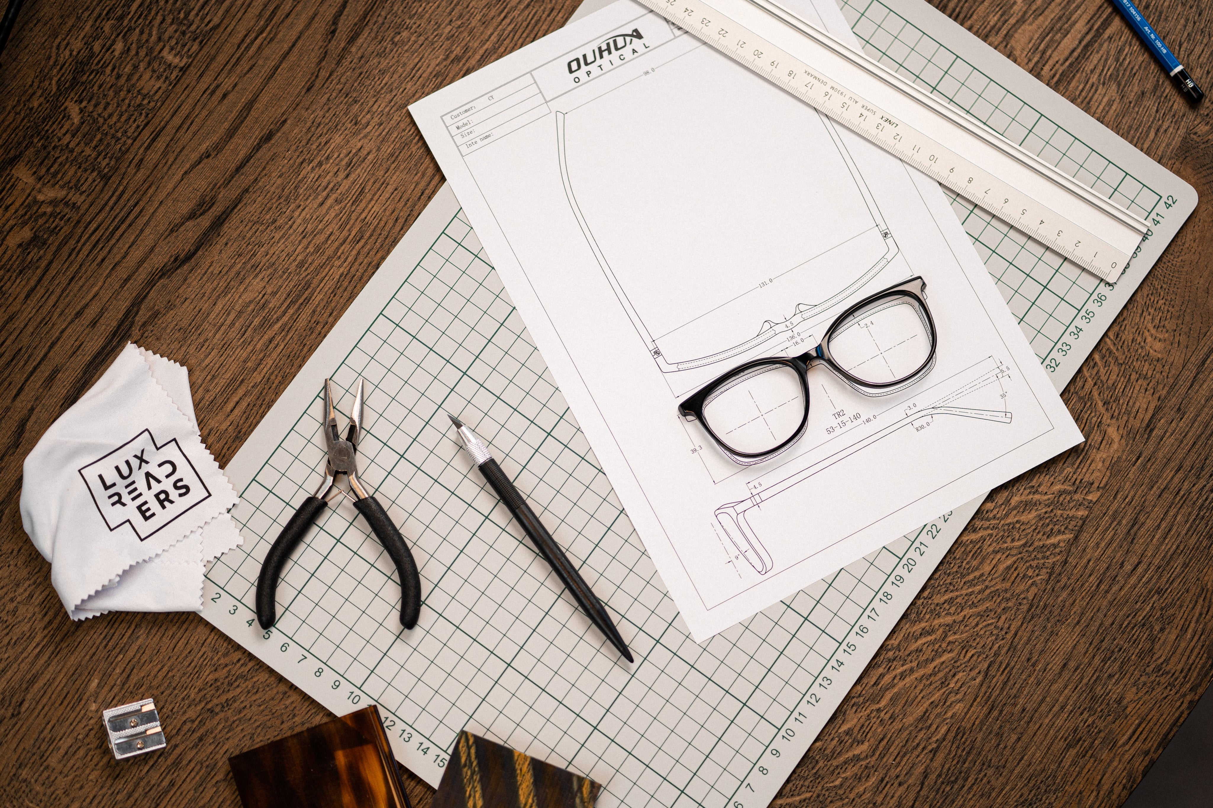 Glasses design sketch with tools, Luxreaders cloth, pliers, and ruler on a wooden table.